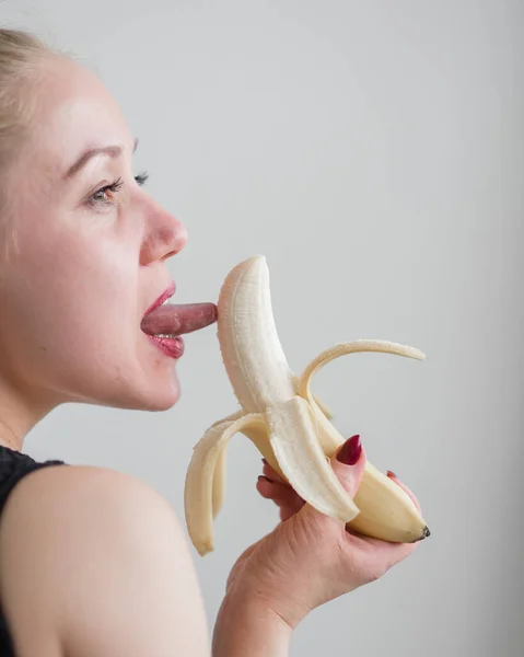 Blondinen i svarta spetsunderkläder äter erotiskt en banan. Attraktiv kvinna med sensuella röda läppar suger sexuellt och slickar en banan. Oral njutning. — Stockfoto