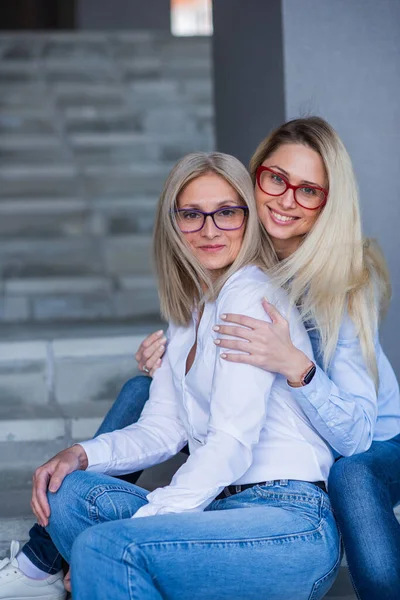 Een mooie blonde met bril en haar volwassen moeder zitten buiten op de trap. Een goed verzorgde oudere vrouw knuffelt haar volwassen dochter stevig. Liefde en familiewaarden. Moederdag. — Stockfoto