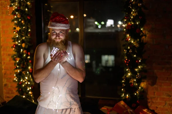 Santa mala. Un hombre barbudo con una camiseta blanca y un sombrero rojo de Santa Claus sonríe slyly y sostiene una guirnalda de Navidad en sus manos . —  Fotos de Stock