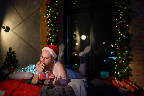 Humour sur le sujet de Noël. Un geek avec une fille aux cheveux roux dans un chapeau du Père Noël se trouve sur un lit et découvre un cadeau. Père Noël a reçu un cadeau . — Photo