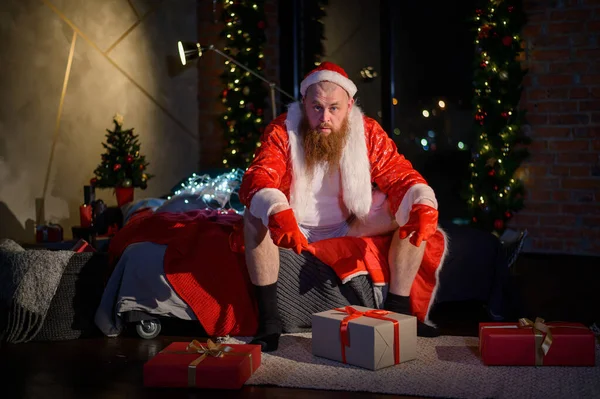 Een slechte slechte Kerstman zit op het bed en pakt cadeautjes in. Vreemde Kerstman in een open pak en wit T-shirt. Een man met een rode baard in een kerstmanpak. — Stockfoto