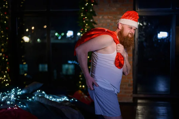 Slechte boze kerstman met een rode baard draagt een tas met geschenken op kerstavond. Een man met een korte broek en een kerstmuts krabt aan zijn kont. Kerstman voor het nieuwe jaar. — Stockfoto