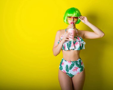 Şarlatanın altında parlak yeşil peruklu ve sarı arka planda poz veren güzel bir genç kadın. Şehvetli dudakları olan çekici bir kız kamerada flört ediyor, pipetle içki içiyor..