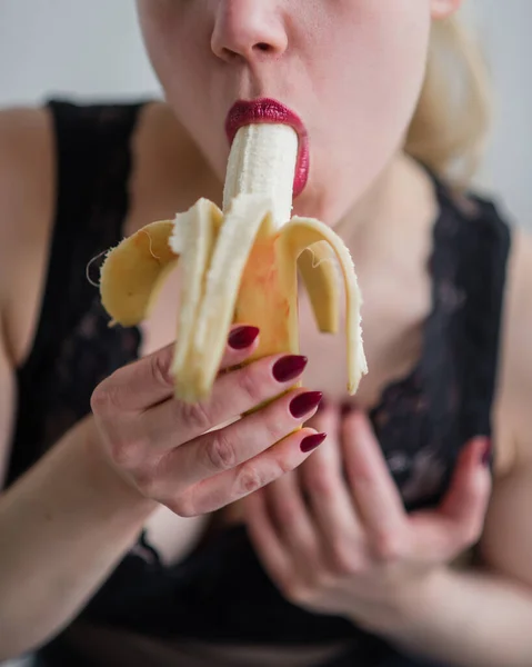 Blondinen i svarta spetsunderkläder äter erotiskt en banan. Attraktiv kvinna med sensuella röda läppar suger sexuellt och slickar en banan. Muntligt nöje. Närbild. — Stockfoto