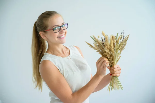 Die Blondine in Brille und weißer Bluse sammelt in einem weißen Atelier einen Strauß getrockneter Blumen und Baumwolle. eine charmante Floristin lächelt bei der Arbeit. — Stockfoto