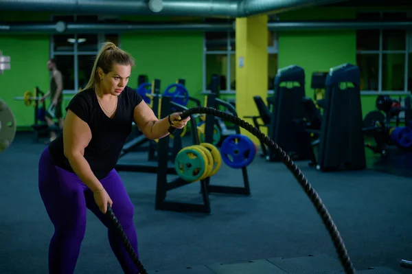 Fat woman doing strength training using battle ropes in the gym. An obese athlete moves the ropes in a wave motion to train fat burning.