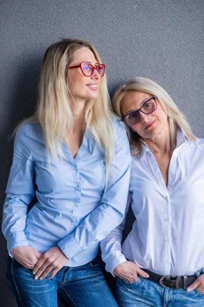 Portrét dvou krásných žen různých generací proti šedé zdi. Rodinná podoba. Matka a dcera v džínové košili a brýlích. Dobře upravená starší žena. — Stock fotografie