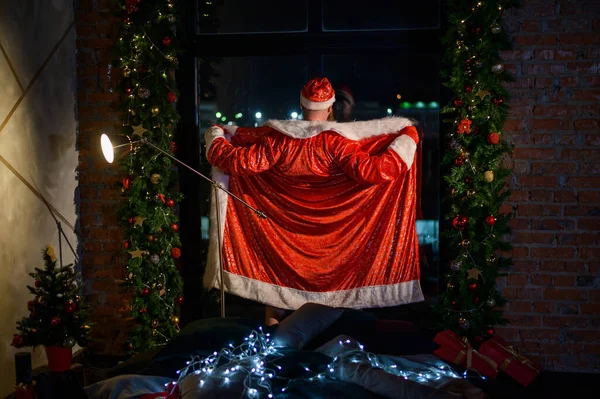 Humorous image of a man in a suit of Santa Claus showing striptease at the window. Bad Santa Claus opened his suit for a joke. Vulgar humor — Stock Photo, Image