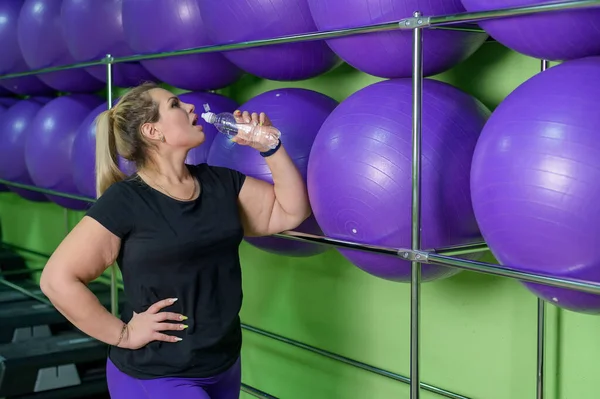 Una donna sovrappeso beve acqua durante un allenamento in palestra. Palle di fitness sullo sfondo . — Foto Stock