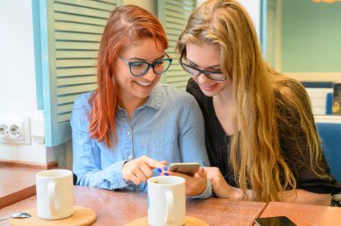 İki heyecanlı genç kız cep telefonu kullanıyor, bir kafede oturuyor ve parmakla işaret ediyor. Gözlüklü kızıl saçlı kadın akıllı telefon kız arkadaşının komik fotoğraflarını gösteriyor. Kahve içerler..