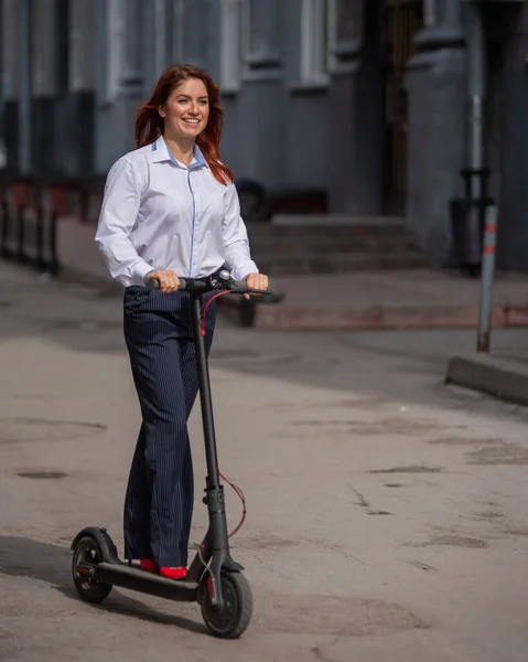 Ein rothaariges Mädchen im weißen Hemd fährt mit einem Elektroroller die Wand entlang. Eine Geschäftsfrau im Hosenanzug und roten High Heels fährt in einem modernen Auto durch die Stadt. Kleiderordnung im Büro. — Stockfoto