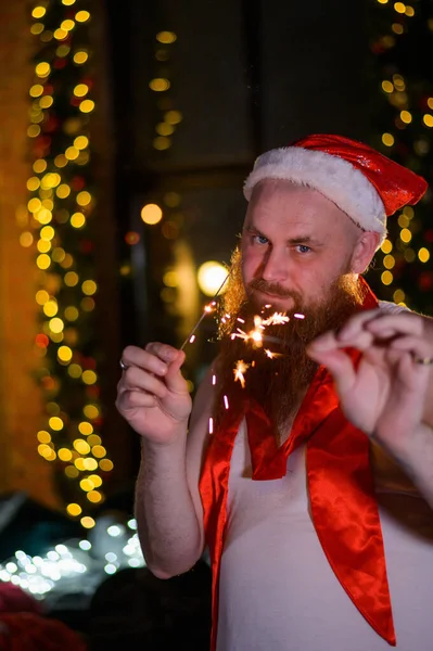 Tomten med julglitter. En man med rött skägg i en jultomte hatt skämtar gör ett glamoröst foto. — Stockfoto