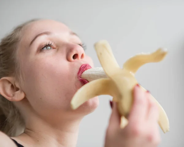Die Blondine in schwarzer Spitzenunterwäsche isst erotisch eine Banane. Attraktive Frau mit sinnlichen roten Lippen saugt und leckt sexuell eine Banane. Mündliches Vergnügen. — Stockfoto