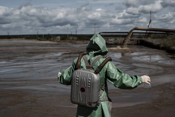 Mužský ekolog v zeleném ochranném obleku a plynové masce odebere vzorek vody ve znečištěném jezeře. Odpad z výroby. Vědec provádí toxikologickou studii toxického odpadu. — Stock fotografie