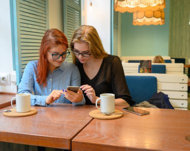 İki heyecanlı genç kız cep telefonu kullanıyor, bir kafede oturuyor ve parmakla işaret ediyor. Gözlüklü kızıl saçlı kadın akıllı telefon kız arkadaşının komik fotoğraflarını gösteriyor. Kahve içerler..
