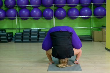 Şişman kadın spor salonunda pilates yapıyor. Yoganın yardımıyla kilo vermeye çalışan aşırı kilolu bir kız kuzgun pozunda duruyor. Sarışın bokasana çalıyor..