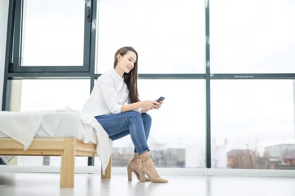 Μια νεαρή γυναίκα με λευκό πουκάμισο μιλάει στο τηλέφωνο κοντά σε ένα μεγάλο παράθυρο σε ένα ξενοδοχείο. Όμορφη κοπέλα διαπραγματεύεται ενώ κάθεται στο κρεβάτι. Εργασία από το σπίτι. — Φωτογραφία Αρχείου
