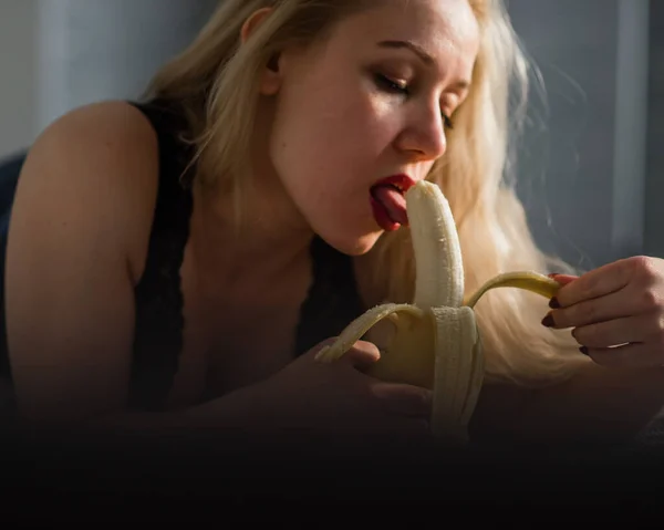 Blondinen i svarta spetsunderkläder äter erotiskt en banan. Attraktiv kvinna med sensuella röda läppar suger sexuellt och slickar en banan. Oral njutning. — Stockfoto