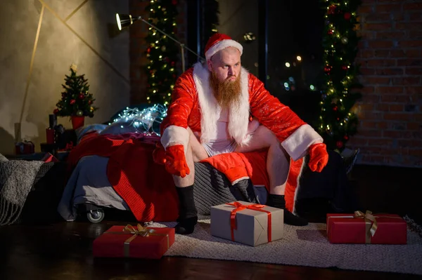 Zły zły Święty Mikołaj z czerwoną brodą siedzi na łóżku i zbiera prezenty w Wigilię. Człowiek w garniturze Świętego Mikołaja przed Nowym Rokiem. — Zdjęcie stockowe