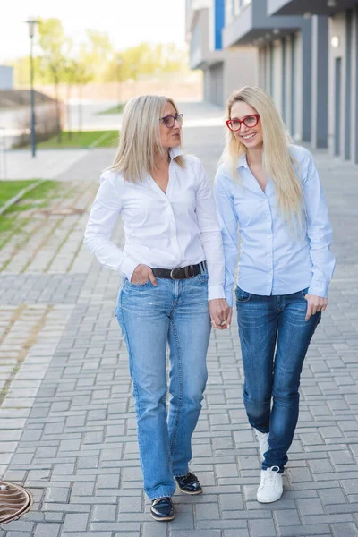 Een mooie blonde met bril en haar oudere moeder lopen samen. Moederdag. Mooie oude vrouw is een oudere burger en haar volwassen dochter. Gelukkige vrouwen van verschillende generaties. — Stockfoto