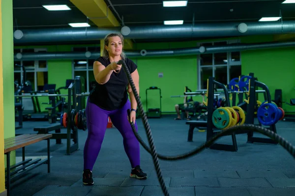 Mujer gorda haciendo entrenamiento de fuerza usando cuerdas de batalla en el gimnasio. La muchacha obesa se ocupa de los ejercicios circulares para la pérdida del peso . — Foto de Stock