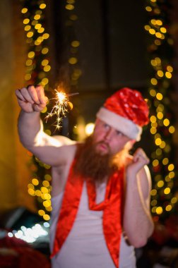 Noel Baba 'nın pırıltıları. Noel Baba şapkalı kızıl sakallı ve alkolik tişörtlü bir adam maytap yakıyor. Parodi.