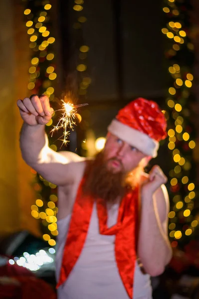 Julemand med jule glimmer. En mand med rødt skæg i julemandens hat og en T-shirt til en alkoholiker brænder stjernekastere. Parodi. - Stock-foto