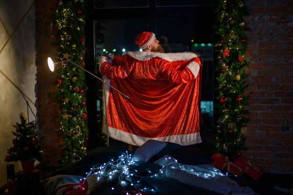 Bad evil Santa Claus opened his robe in front of the window on Christmas Eve. A man in a suit of Santa Claus shows a striptease in the New Year. Exhibitionist on holidays. — Stock Photo, Image
