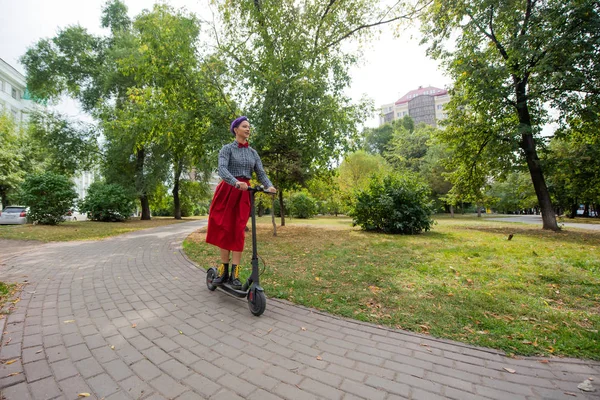 Una joven con el pelo morado monta una scooter eléctrica en un parque. Una chica elegante con un templo afeitado en una camisa a cuadros, una falda roja larga y una corbata de lazo está montando alrededor de la ciudad en un dispositivo moderno . — Foto de Stock