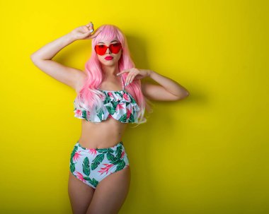 Pembe peruklu ve sarı arka planda poz veren renkli bikinili güzel kız. Yapay uzun saçlı ve kalp şeklinde pembe gözlüklü bir kadın..