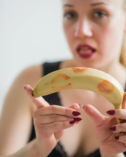 A szőkének van egy hámozatlan banánja vörös rúzsnyomokkal. Az orális szex fogalma. Csábító nézés. Étvágygerjesztő gyümölcs. — Stock Fotó