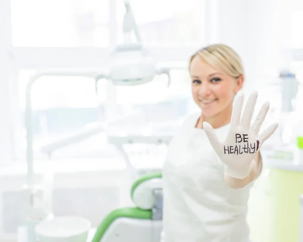 Amistosa dentista femenina sonriendo y dando choca esos cinco. Médico rubio con guantes en un consultorio en un centro médico. La inscripción en la mano ser saludable . —  Fotos de Stock