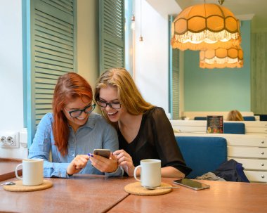 İki heyecanlı genç kız cep telefonu kullanıyor, bir kafede oturuyor ve parmakla işaret ediyor. Gözlüklü kızıl saçlı kadın akıllı telefon kız arkadaşının komik fotoğraflarını gösteriyor. Kahve içerler..