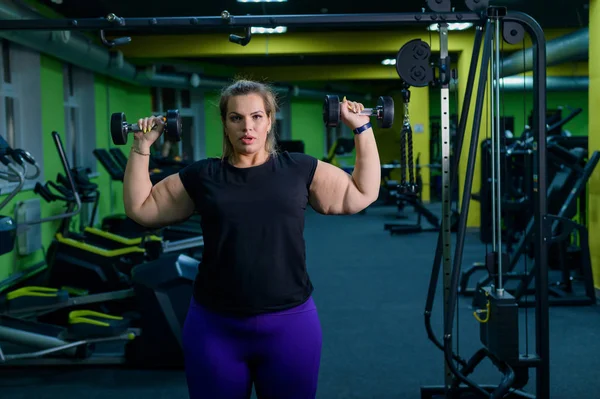 An obese woman is working on losing weight. Fat blondes train biceps with a dumbbell in the trainer room. A lot of excess weight due to poor eating habits and lack of self-control.