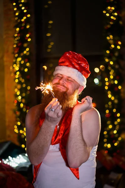 Télapó karácsonyi csillogással. Egy vörös szakállú férfi a Mikulás kalapjában és egy alkoholista pólója csillagszórókat éget. Paródia. — Stock Fotó