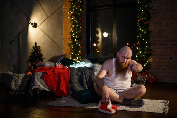 Imagen humorística de un hombre sin pantalones delante de Navidad. El calvo Papá Noel con barba roja desempaqueta un regalo. Una parodia de una sesión de fotos glamurosa de Año Nuevo para mujeres. Broma. . — Foto de Stock