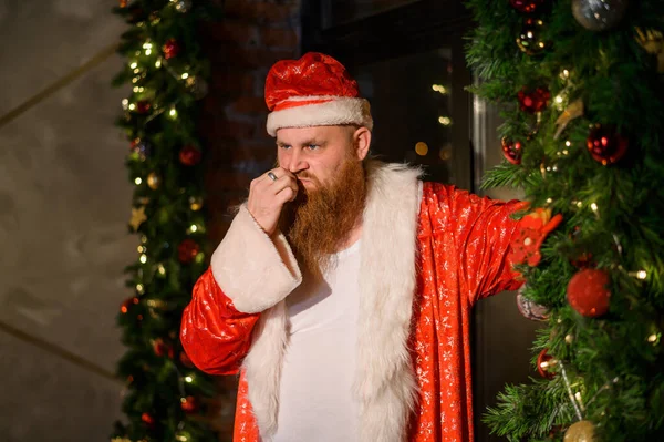 Bad Santa claus einsam an Weihnachten. Aggressiver Mann im Weihnachtsmann-Anzug auf dem Hintergrund von Silvesterdekorationen. — Stockfoto