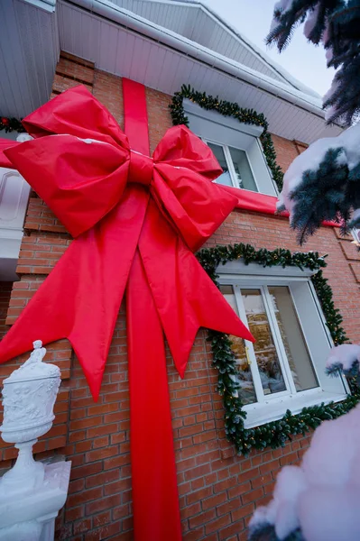 A large bow from a red ribbon on the facade of a brick country house. Christmas decoration with coniferous trees on the window. — Stockfoto