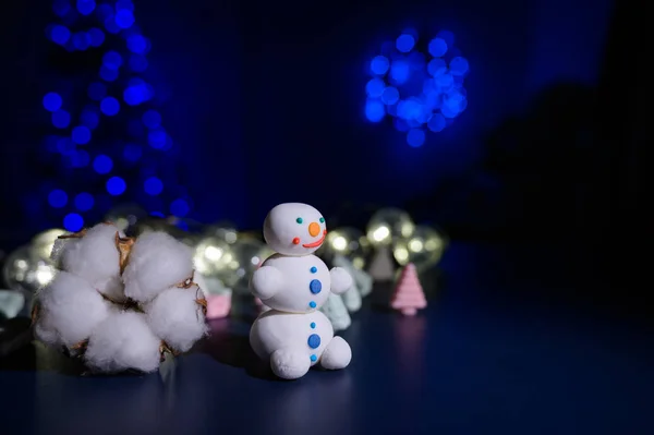 Сладкий снеговик из кондитерской мастики на фоне зефира и белой кружки с какао. Свитера и бутон хлопок в темноте на фоне елки в огнях . — стоковое фото