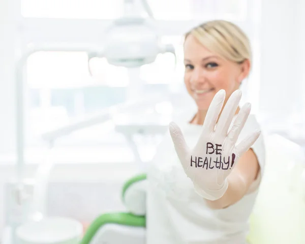 Amichevole dentista femminile sorridente e dando il cinque. Medico biondo con i guanti in un ufficio in un centro medico. L'iscrizione sulla mano essere sano . — Foto Stock
