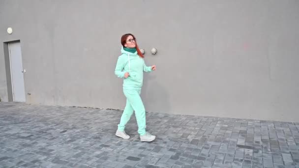 Una mujer en un cálido traje deportivo de menta y bufanda está caminando por la calle y bailando. Una chica pelirroja feliz con una sudadera azul, pantalones y gafas camina y se divierte. Temprano soleado mañana helada. — Vídeos de Stock