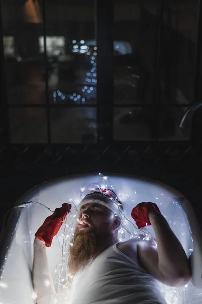 Un'immagine umoristica di un Babbo Natale ubriaco sdraiato in un bagno in mutande con le luci di Natale. Un uomo con la barba rossa in un cappello di Babbo Natale tiene in mano una ghirlanda. vista dall'alto . — Foto Stock