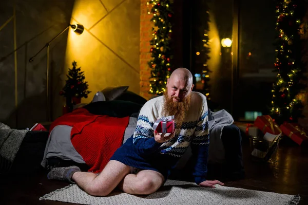Un hombre con una larga barba roja se sienta en el suelo sin pantalones en un suéter de invierno. hombre está sosteniendo un regalo envuelto en el fondo de las decoraciones de Año Nuevo y luces. Árbol de Navidad. Parodia, humor. —  Fotos de Stock