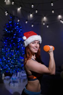 Noel Baba şapkalı güzel kızıl atlet pazı egzersizi yapıyor. Spor kıyafetli kadın, yılbaşı ağacının üzerinde karanlık bir stüdyoda elinde dambıllar tutuyor..