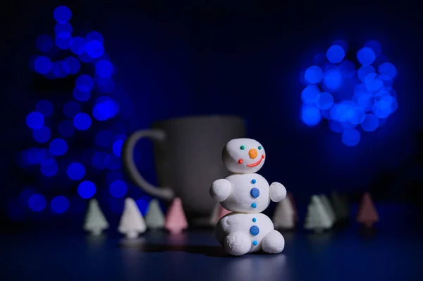 Крупный план сладкого снеговика из кондитерской мастики на фоне зефира и белой кружки с какао. Конфеты в темноте на фоне рождественской елки в огнях . — стоковое фото