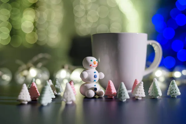 Cute snowman and little Christmas trees made of marshmallows on the background of Christmas lights. White mug of hot chocolate and sweets. — Stock Photo, Image