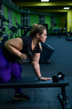 Obez bir kadın spor salonundaki bir bankta dambıllarla antrenman yapıyor. Şişko sarışın fitness sayesinde kilo veriyor..