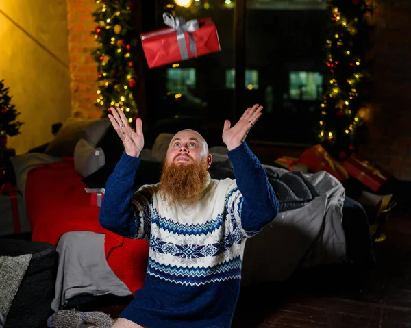 Muž s dlouhým rudým vousem sedí na podlaze bez kalhot v zimním svetru. muž drží zabalený dárek na pozadí novoroční výzdoby a světla. Vánoční stromek. Parody, humor. — Stock fotografie