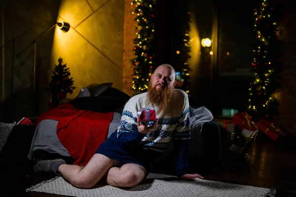 A man with a long red beard sits on the floor without pants in a winter sweater. man is holding a wrapped present on the background of New Year decorations and lights. Christmas tree. Parody, humor. — Stock Photo, Image