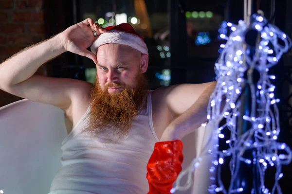 Humorvol beeld van een slechte kerstman die een bad neemt met kerstverlichting. Een man met een rode baard in een kerstman hoed en ondergoed houdt een bloemenslinger in zijn handen.. — Stockfoto